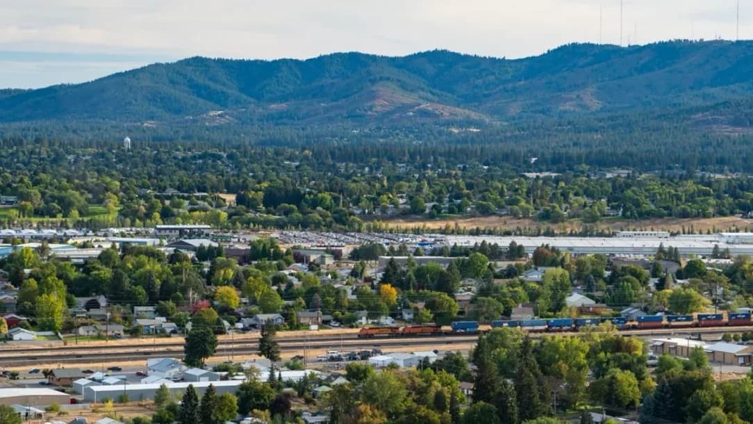 Spokane Valley seen from above — homes and industrial buildings across the valley floor, a rail yard and freight train in the foreground, forested hills behind.