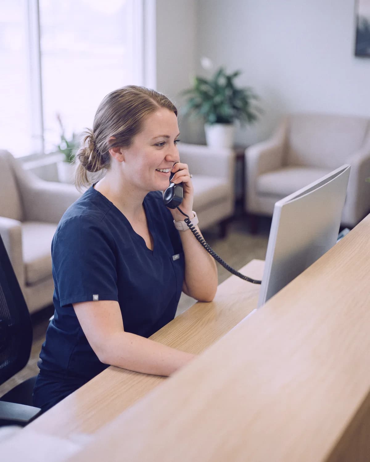 A dental office manager at the front desk, smiling on the phone while she works at her computer, the waiting room behind her.
