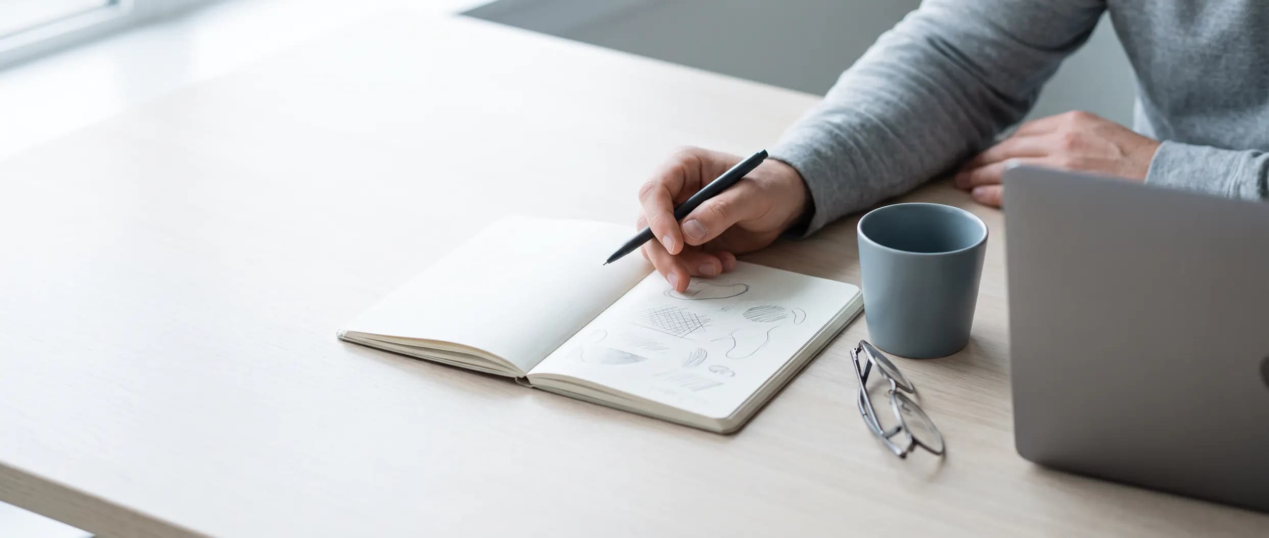 An open notebook of hand-sketched page ideas on a desk beside a laptop and a coffee — content planned before it is written.