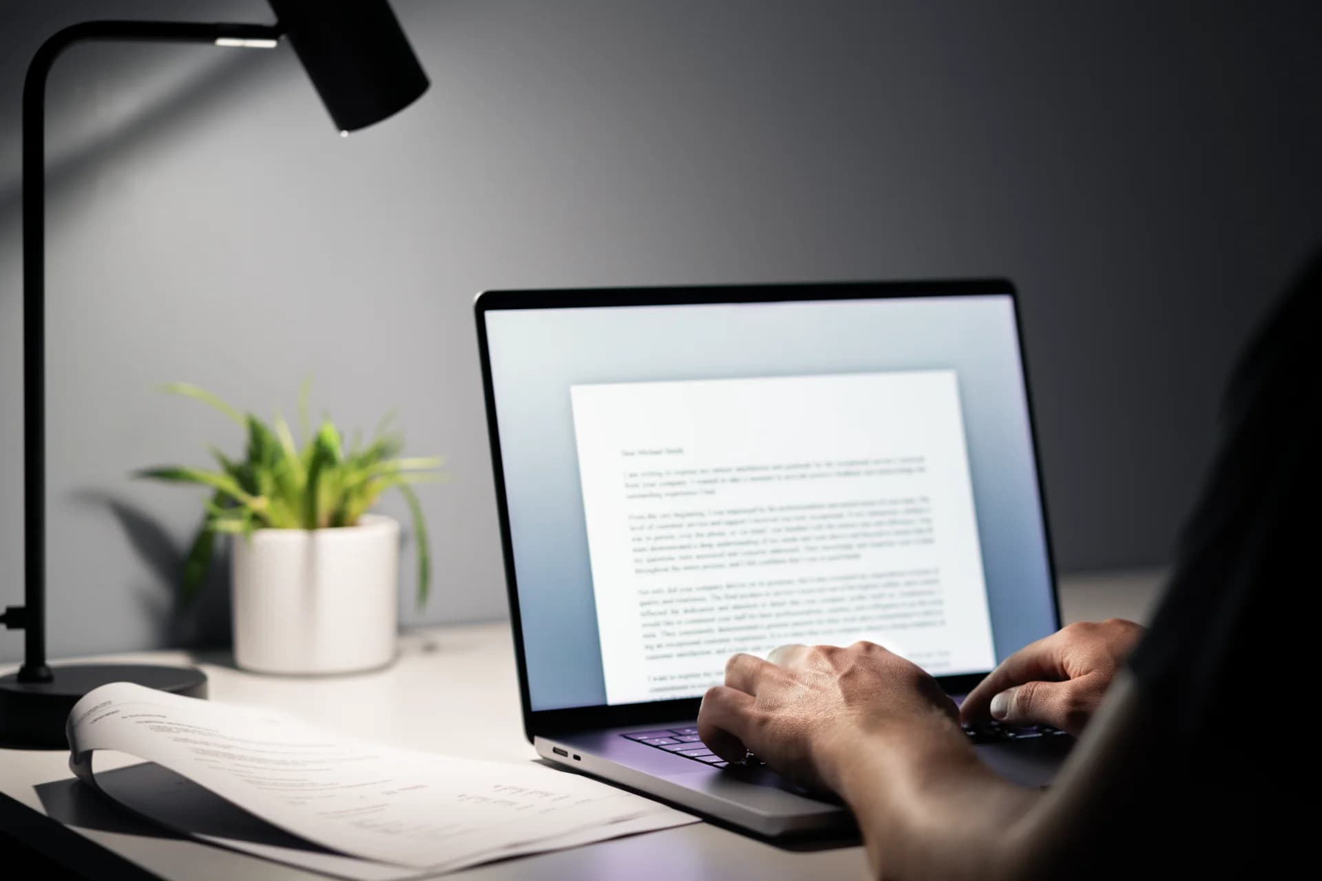 A writer at a laptop under a desk lamp, drafting a document on screen with a printed page laid out on the desk beside it.
