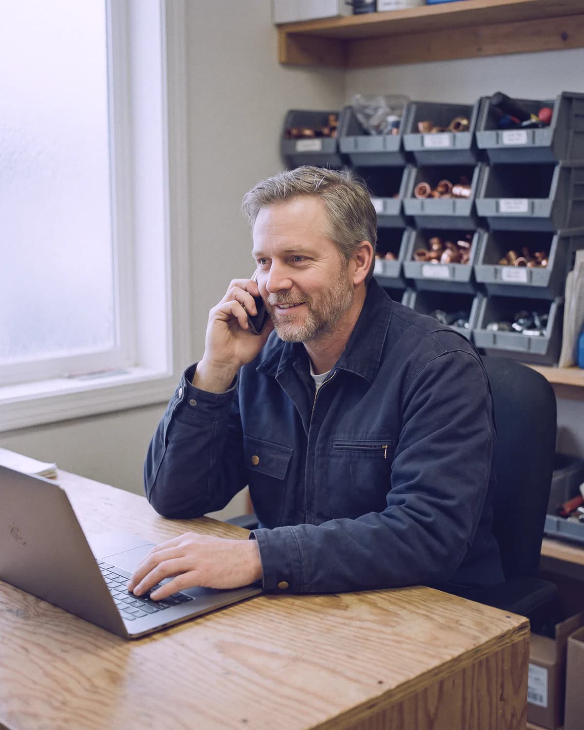 The owner of a plumbing and heating company at his office desk, smiling on the phone while he works on his laptop, parts bins on the shelves behind him.