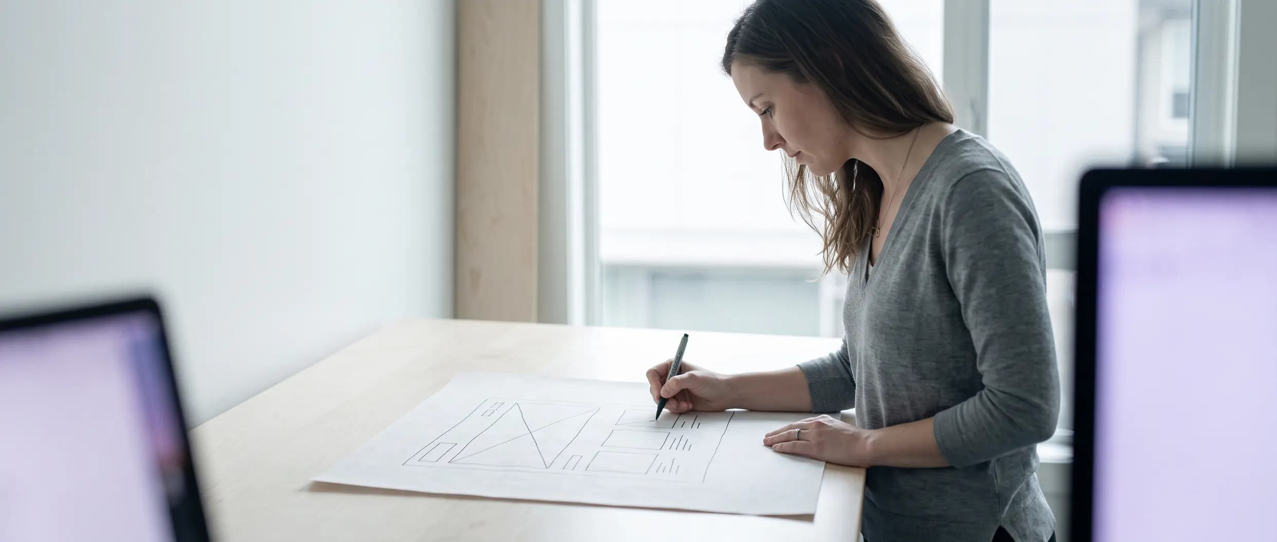 A designer standing at a desk, drawing a page-layout wireframe on paper before a site gets rebuilt.